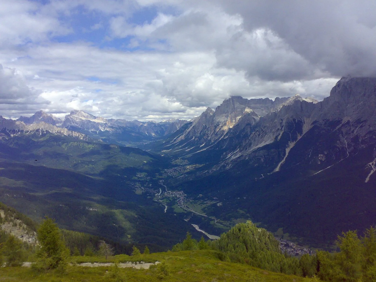 Panorama estivo della Val del Boite vista dall'alto, con il fondovalle verde, i paesi e le pareti dolomitiche sotto un cielo di nuvole